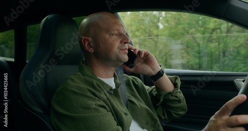 A man is talking on a mobile phone while driving a car. He's smiling. View from the car interior. Summer day photo