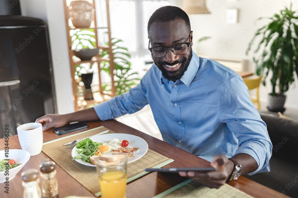 A smiling African-American man enjoys breakfast while using his tablet. He is seated at a table with a healthy meal and juice, in a cozy and modern home setting. A smiling African-American man enjoys breakfast while using his tablet. He is seated at a table with a healthy meal and juice, in a cozy and modern home setting.