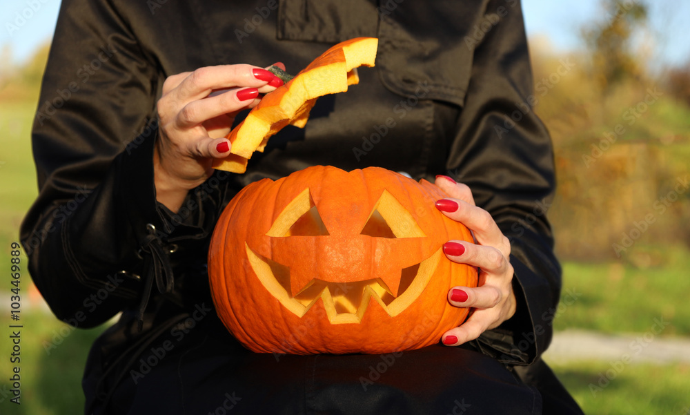 Woman's hands with red blood nail polish, holding orange carved Halloween pumpkin and opening top of jack-o-lantern head. Halloween traditional decoration. Sunny day outdoors.