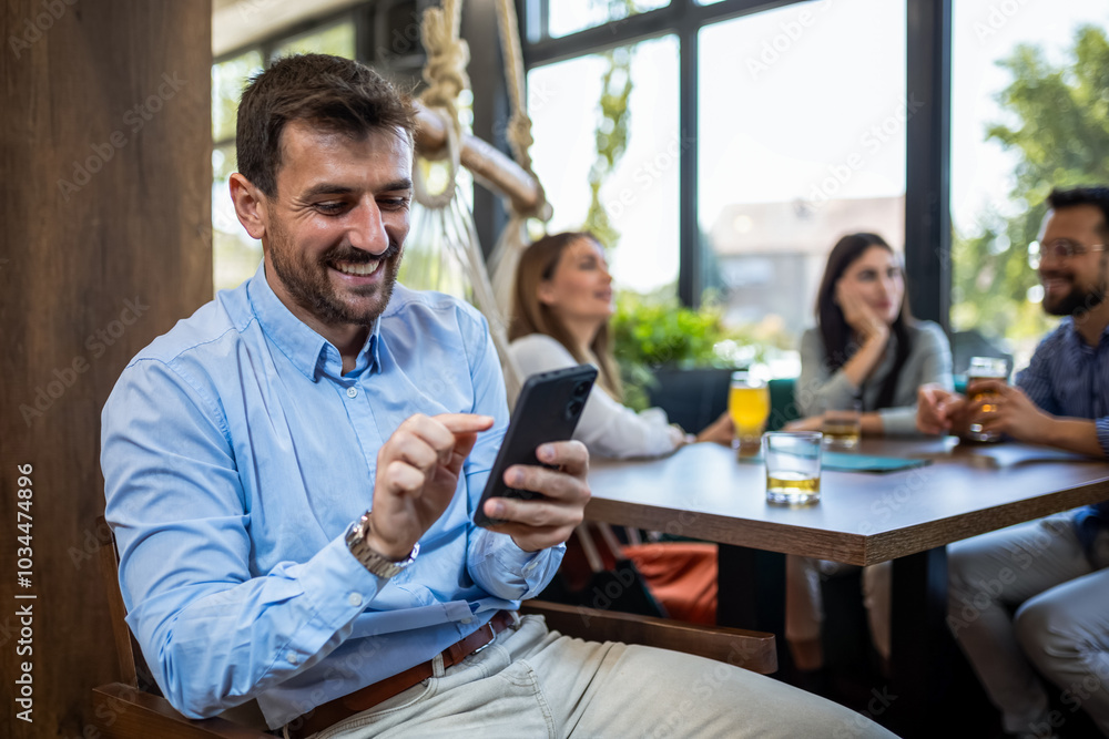 Young businessman using his phone while in restaurant with his friends in the background. Young businessman using his phone while in restaurant with his friends in the background.