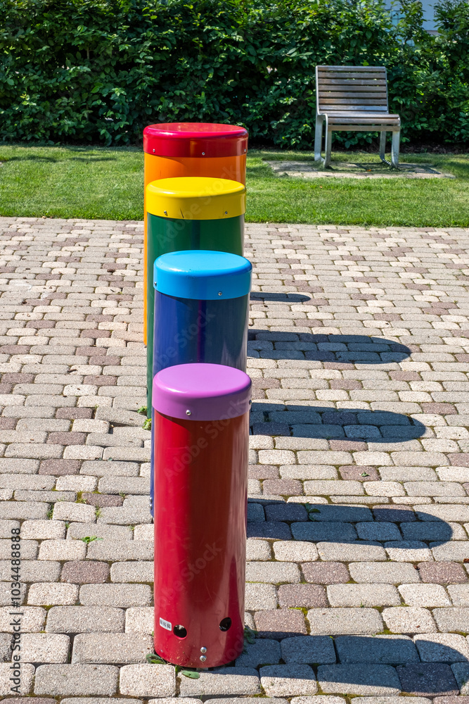Brightly colored cylindrical objects arranged in a park setting next to a bench on a sunny day Brightly colored cylindrical objects arranged in a park setting next to a bench on a sunny day