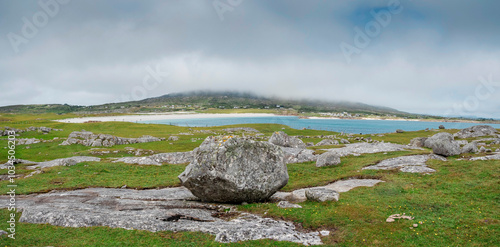 A large rock sits in a grassy field next to a body of water. The sky is cloudy, but the sun is still shining through the clouds. The scene is peaceful and serene, Dogs bay, county Galway, Ireland photo