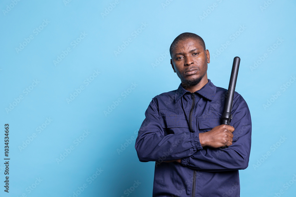 African american security guard feels proud with his baton in studio, posing with confidence with his weapon and professional uniform. Skilled agent standing against blue background. African american security guard feels proud with his baton in studio, posing with confidence with his weapon and professional uniform. Skilled agent standing against blue background.