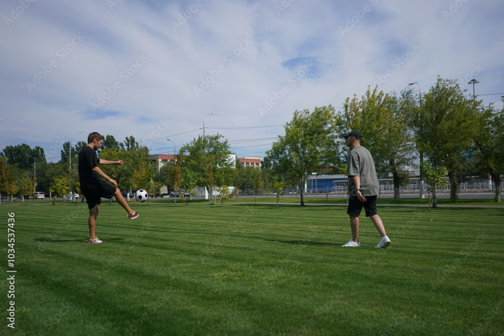 Obraz premium Two men are outdoors in a park, practicing their soccer juggling skills. One man performs ball tricks while the other watches, enjoying the sunny day among the trees