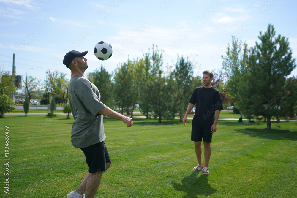Two men are outdoors in a park, practicing their soccer juggling skills. One man performs ball tricks while the other watches, enjoying the sunny day among the trees Two men are outdoors in a park, practicing their soccer juggling skills. One man performs ball tricks while the other watches, enjoying the sunny day among the trees