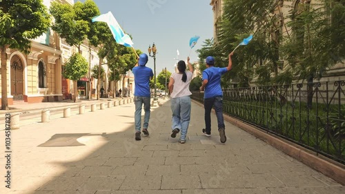 Rear view of a group of three people running through the streets of a Latin American city, celebrating their country's national holidays. They are carrying cloth flags and holding them high.