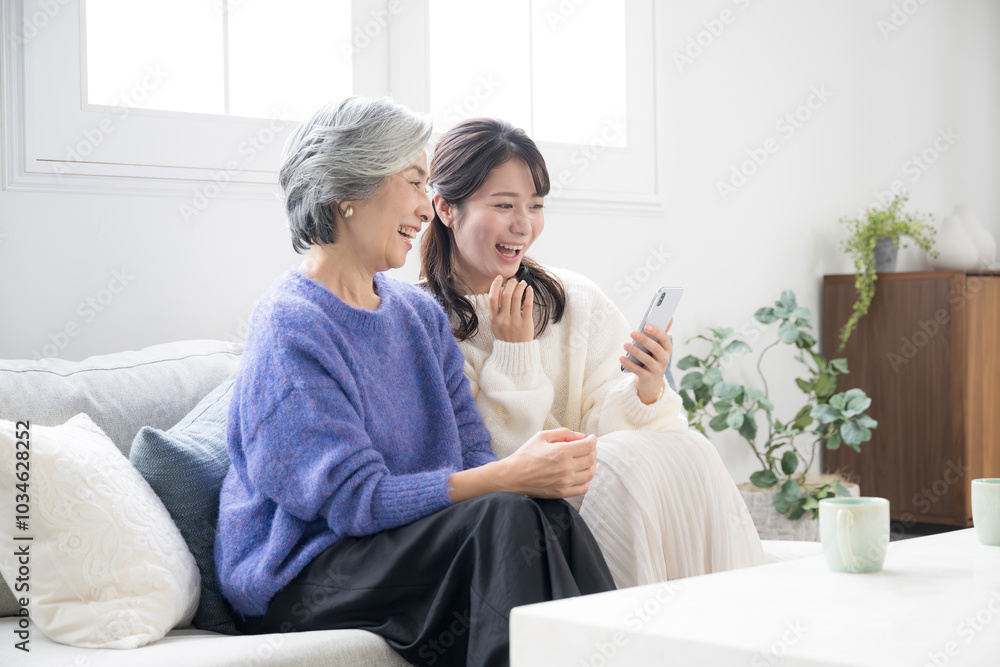 リビングで笑顔で話す、スマホを見ながら過ごす仲良し親子(母と娘)広角 リビングで笑顔で話す、スマホを見ながら過ごす仲良し親子(母と娘)広角