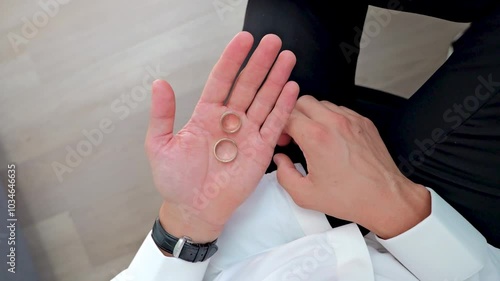 The man sits on a chair around the window and holds two rings in his hand.