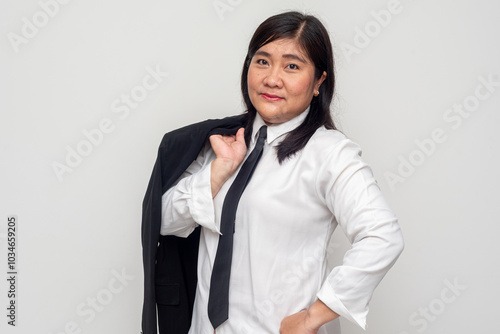 A mature Indonesian woman is seen in a confident pose, wearing a white shirt with a black tie. She holds her black blazer over her shoulder, smiling slightly with a relaxed and professional demeanor photo