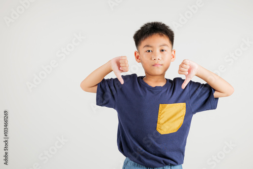 Portrait disheartened Asian young kid boy displaying a thumbs down gesture, symbolizing dislike. Studio shot isolated on white with ample copy space, primary child portraying negative and sad look