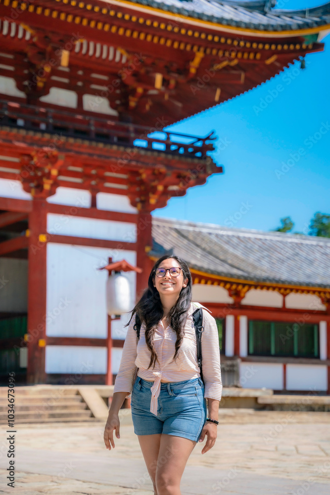 A happy young Latina woman, dressed casually with glasses and a backpack, smiles while exploring the historic Todaiji Kondo Hakkaku Toro in Nara, Japan, on a sunny day. A happy young Latina woman, dressed casually with glasses and a backpack, smiles while exploring the historic Todaiji Kondo Hakkaku Toro in Nara, Japan, on a sunny day.