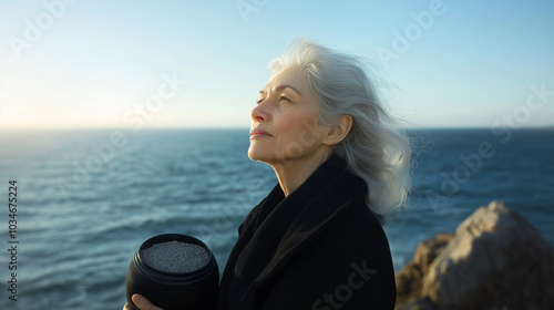Elderly woman in black holds an urn with ashes by the ocean at sunrise