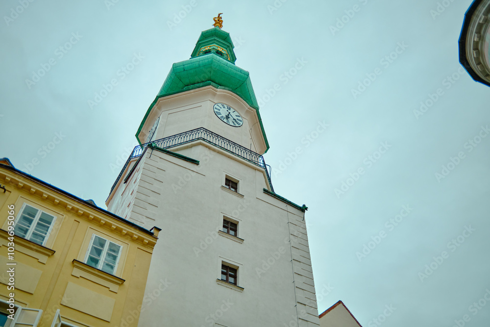 Photograph of a historic building in Bratislava, Slovakia, showcasing a distinctive green tower with a clock and red-tiled roof. Photograph of a historic building in Bratislava, Slovakia, showcasing a distinctive green tower with a clock and red-tiled roof.