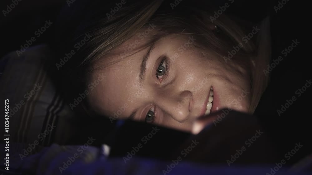 Young woman smiling while using smartphone in bed at night. The light from the phone illuminates her face. Young woman smiling while using smartphone in bed at night. The light from the phone illuminates her face.
