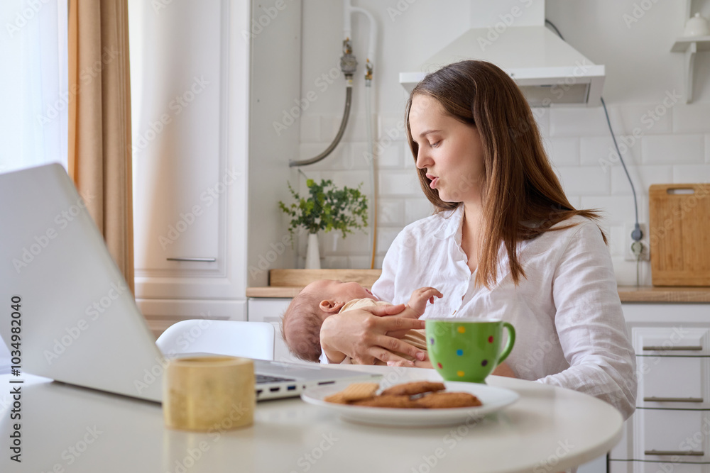 Brown haired woman being busy working remotely from home on her laptop in kitchen calming down her newborn baby doing her business tasks online and caring for her infant child Brown haired woman being busy working remotely from home on her laptop in kitchen calming down her newborn baby doing her business tasks online and caring for her infant child
