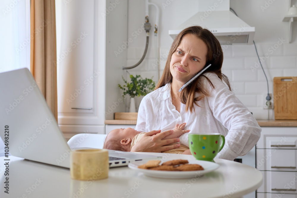 Displeased woman handling business call on her smartphone while working on her laptop in kitchen and looking after her newborn baby Displeased woman handling business call on her smartphone while working on her laptop in kitchen and looking after her newborn baby