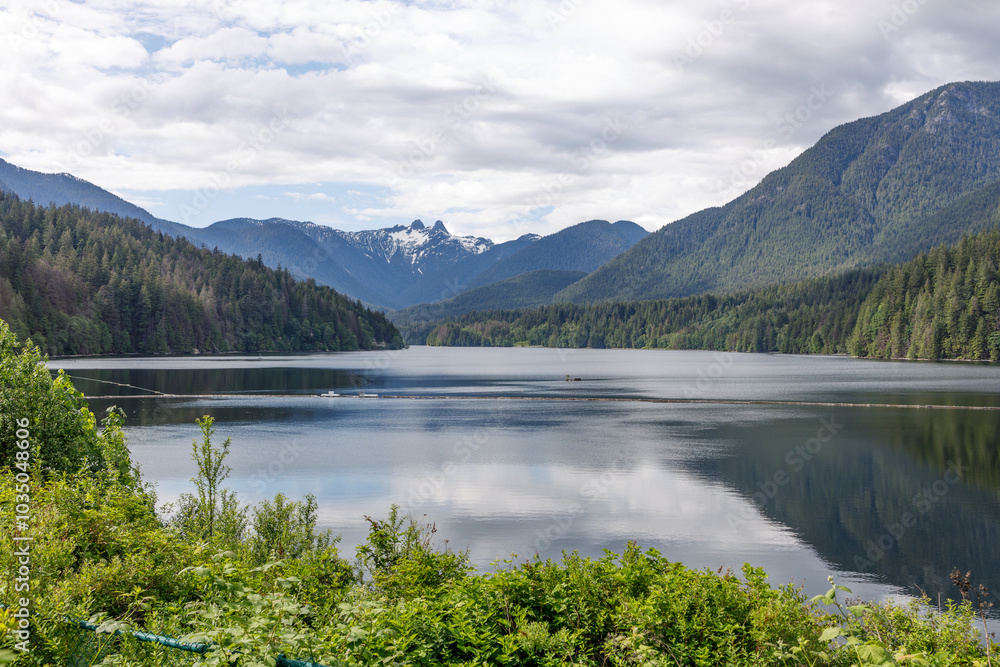 A serene view of Capilano Reservoir surrounded by dense forest, with the majestic Lions Peaks in the background A serene view of Capilano Reservoir surrounded by dense forest, with the majestic Lions Peaks in the background