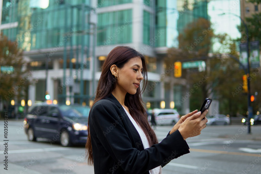Fototapeta premium Hispanic businesswoman using mobile phone on a city street with tall buildings in the background