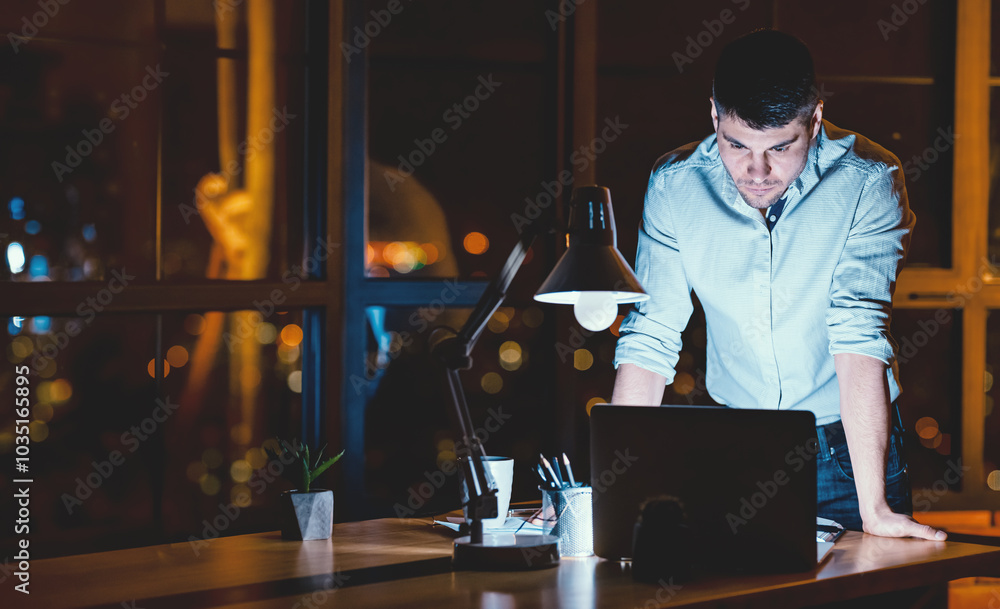 Young Entrepreneur Guy Standing Working On Laptop In Modern Office At Night. Empty Space, Low Light Young Entrepreneur Guy Standing Working On Laptop In Modern Office At Night. Empty Space, Low Light
