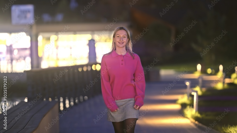Fototapeta premium A young woman leisurely strolls along a beautifully illuminated waterfront path during the enchanting evening