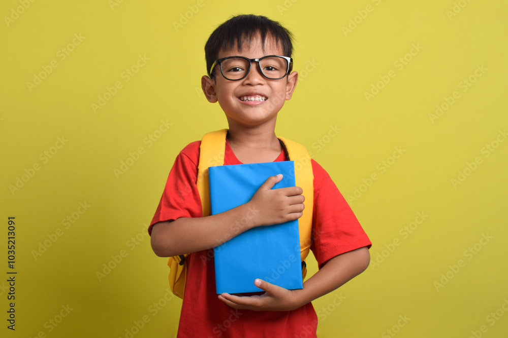 A smiling Asian boy holding a book isolated on a yellow background. A little boy getting ready for school A smiling Asian boy holding a book isolated on a yellow background. A little boy getting ready for school