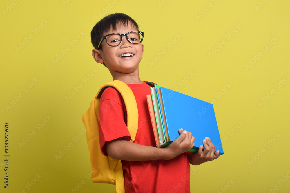 Portrait of Asian boy wearing backpack carrying many books for study isolated on yellow background. Back to school. Education concept Portrait of Asian boy wearing backpack carrying many books for study isolated on yellow background. Back to school. Education concept