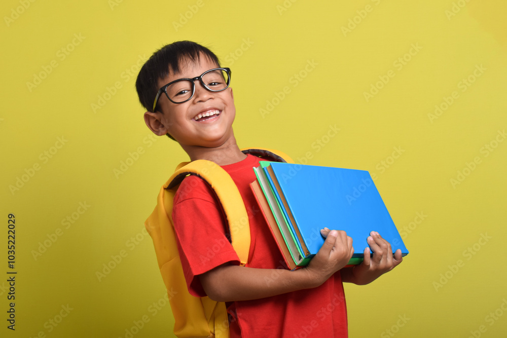 Portrait of Asian boy wearing backpack carrying many books for study isolated on yellow background. Back to school. Education concept Portrait of Asian boy wearing backpack carrying many books for study isolated on yellow background. Back to school. Education concept