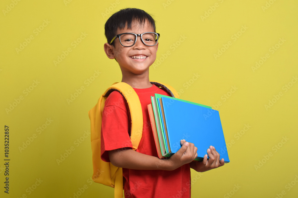 Portrait of Asian boy wearing backpack carrying many books for study isolated on yellow background. Back to school. Education concept Portrait of Asian boy wearing backpack carrying many books for study isolated on yellow background. Back to school. Education concept
