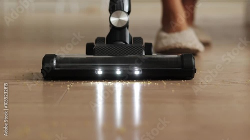 Housekeeping home. Close up unrecognisable woman is using wireless vacuum cleaner with illumination for cleaning kitchen garbage, loose groats from laminate floor. Modern household appliances.