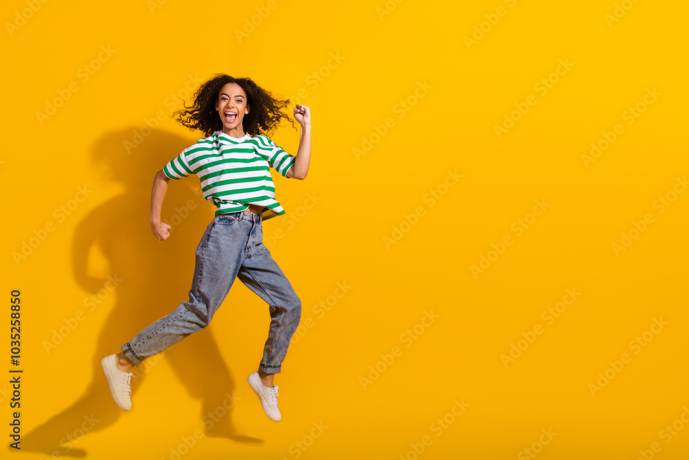Full body portrait of nice young girl jump raise fist empty space wear striped t-shirt isolated on yellow color background Full body portrait of nice young girl jump raise fist empty space wear striped t-shirt isolated on yellow color background