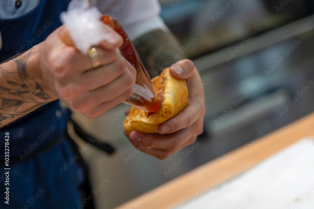 Close-up side view of cook filling fresh baked croissant with fruit or berry jam using icing bag in professional bakery kitchen. Soft focus. Pastry preparation theme. Close-up side view of cook filling fresh baked croissant with fruit or berry jam using icing bag in professional bakery kitchen. Soft focus. Pastry preparation theme.