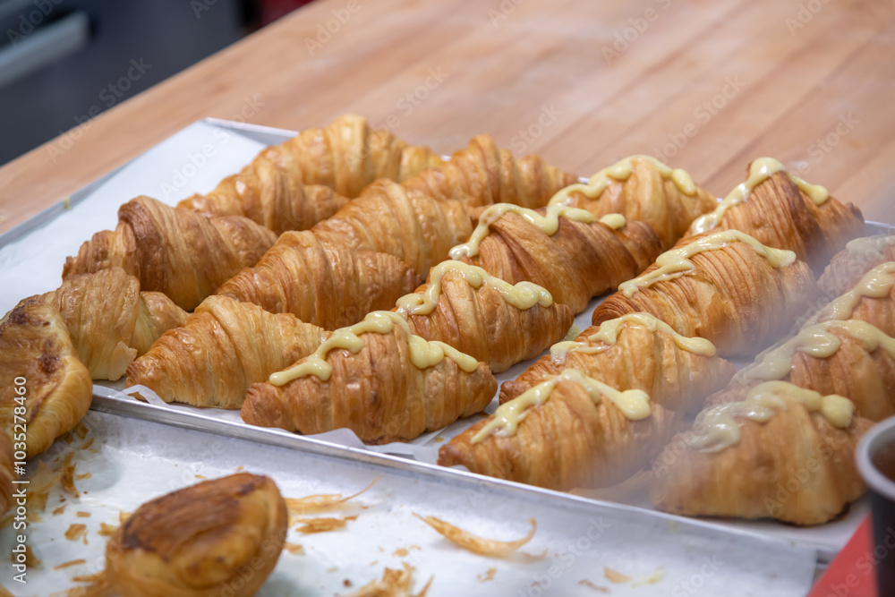 Close-up side view of fresh baked brown croissants lying on white wax paper on baking tray on professional wooden kitchen table. Part of buns covered with custard. Puff pastry preparation theme. Close-up side view of fresh baked brown croissants lying on white wax paper on baking tray on professional wooden kitchen table. Part of buns covered with custard. Puff pastry preparation theme.
