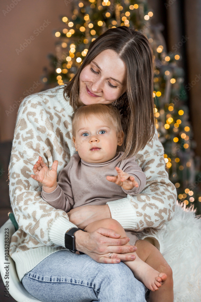 A beautiful little blond boy of 1 year old is sitting with his mother on the background of a Christmas tree. The concept of celebrating Christmas and New Year. A beautiful little blond boy of 1 year old is sitting with his mother on the background of a Christmas tree. The concept of celebrating Christmas and New Year.