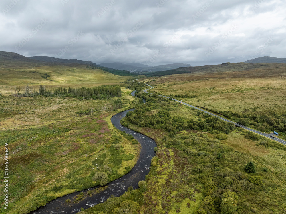 Aerial view of winding road and serene river in lush green landscape surrounded by majestic mountains, Portree, United Kingdom. Aerial view of winding road and serene river in lush green landscape surrounded by majestic mountains, Portree, United Kingdom.