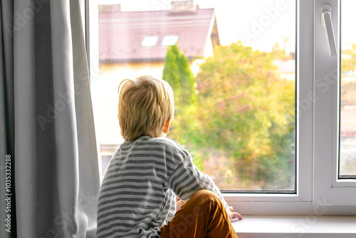 Little boy child kid in pajamas looking sadly through window and wants to go out for a walk and play.Stay at home quarantine,bad weather,punishment
