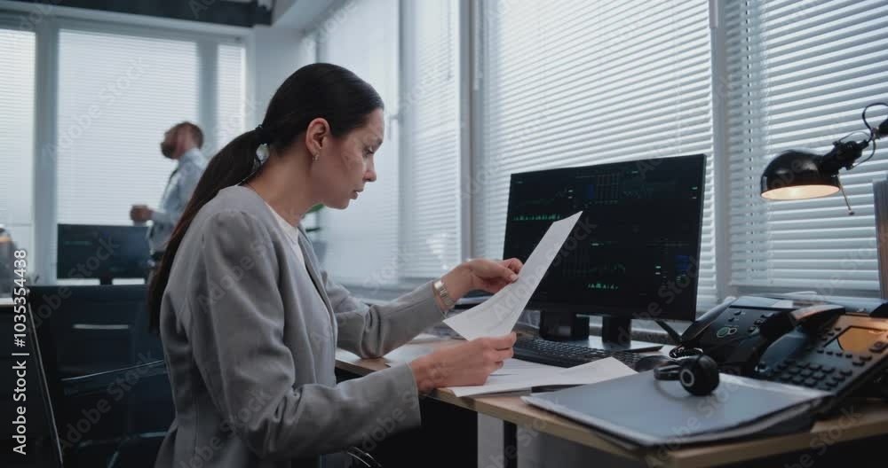 Beautiful adult female financial specialist working with papers, searching and analyzing data, graphs and charts of financial metrics changing on computer monitor. Working in modern business office. Beautiful adult female financial specialist working with papers, searching and analyzing data, graphs and charts of financial metrics changing on computer monitor. Working in modern business office.