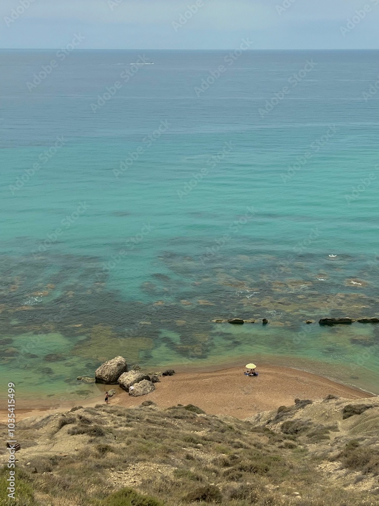 Obraz premium Beach with turquoise waters and rocky shoreline, with beachgoers under an umbrella. Sicily, Italy