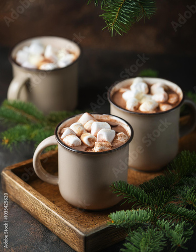 Mugs of creamy hot cocoa topped with marshmallows on wooden tray. Green fir branches.