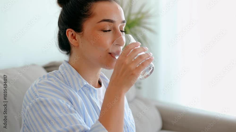 Beautiful happy calm arabian or indian brunette woman in casual wear is drinking a glass cup of clean water in the morning, while sits on sofa in living room at home, leads healthy life style concept Beautiful happy calm arabian or indian brunette woman in casual wear is drinking a glass cup of clean water in the morning, while sits on sofa in living room at home, leads healthy life style concept