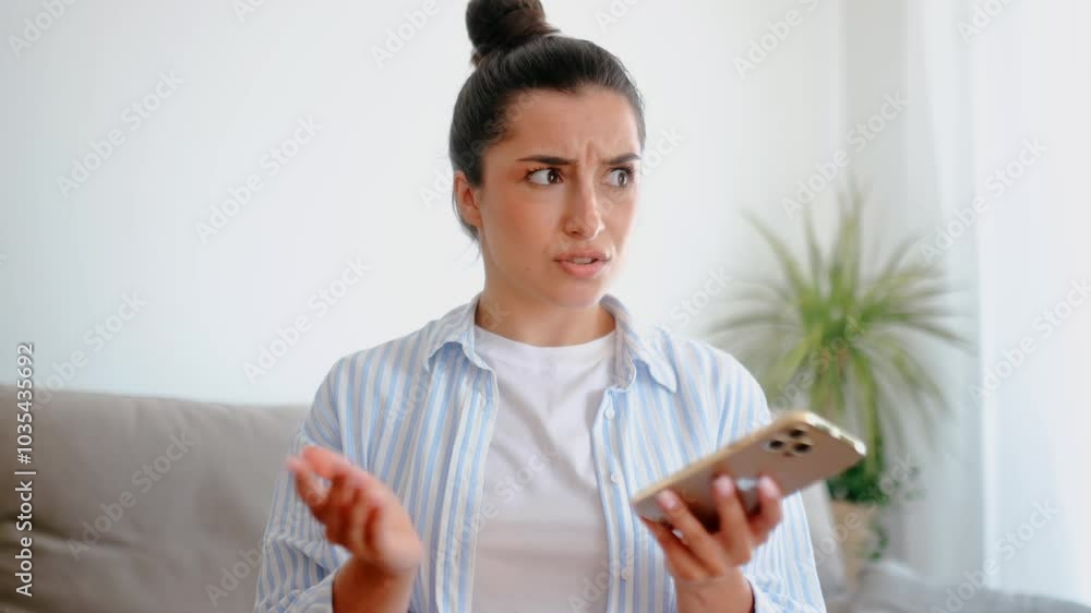 Confused shocked caucasian brunette woman in stylish casual clothes, sitting in the living room, holding her smartphone, looking at puzzled at it, received unexpected news, surprised facial expression Confused shocked caucasian brunette woman in stylish casual clothes, sitting in the living room, holding her smartphone, looking at puzzled at it, received unexpected news, surprised facial expression