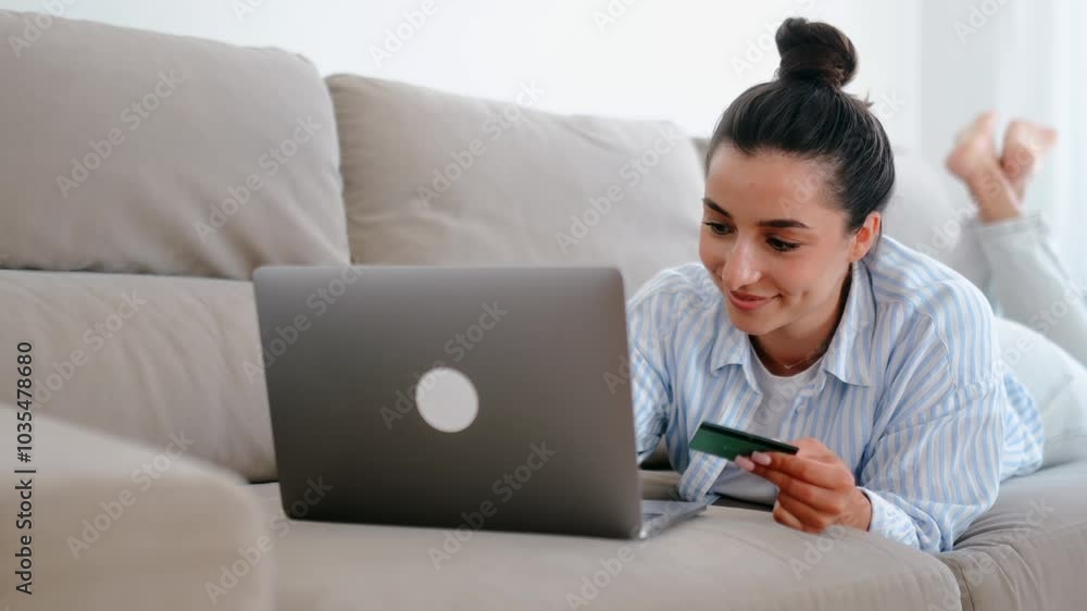 Charming happy caucasian woman in casual clothes lies on a couch in a home living room with a laptop, holding a credit card, making shopping or online banking in mobile app, pay bills, smiles Charming happy caucasian woman in casual clothes lies on a couch in a home living room with a laptop, holding a credit card, making shopping or online banking in mobile app, pay bills, smiles