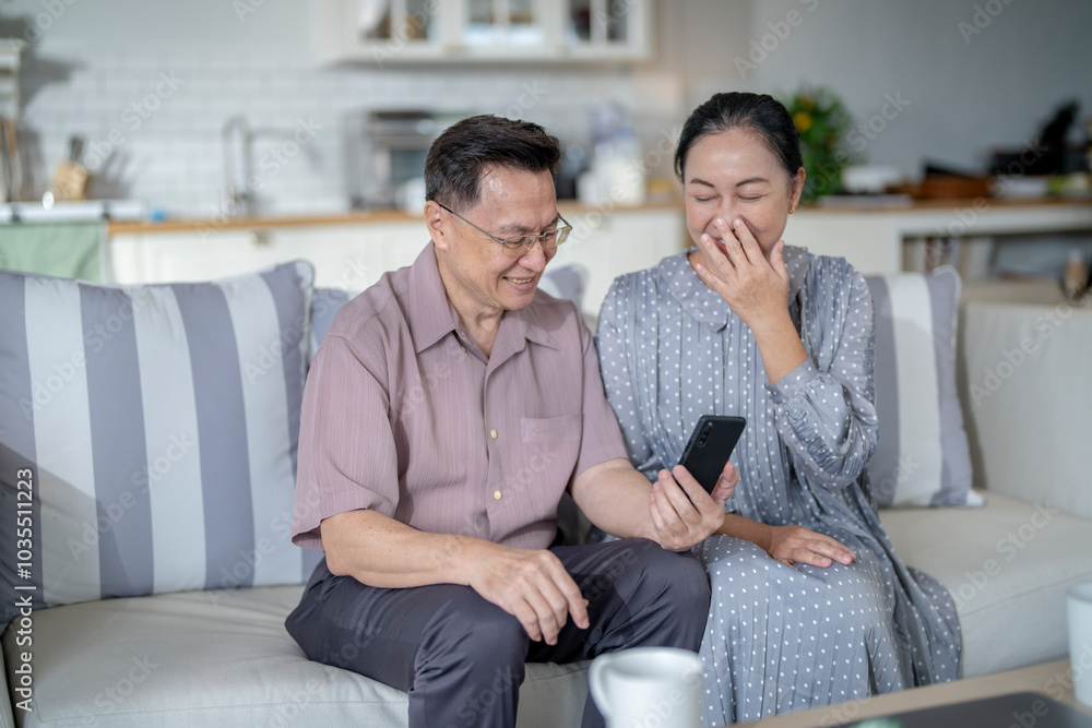 Fototapeta premium An elderly couple sits together in their cozy living room, smiling and enjoying a moment as they look at something on a smartphone. Their warmth and affection capture a peaceful home atmosphere.