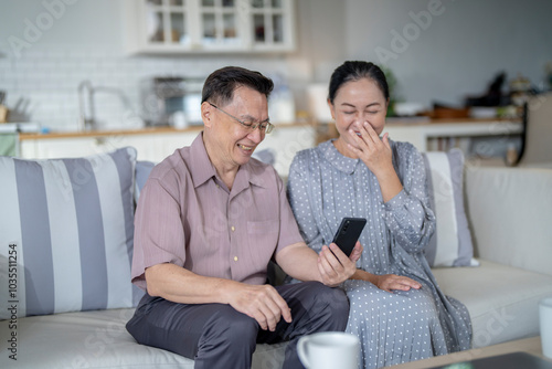 An elderly couple sits together in their cozy living room, smiling and enjoying a moment as they look at something on a smartphone. Their warmth and affection capture a peaceful home atmosphere.