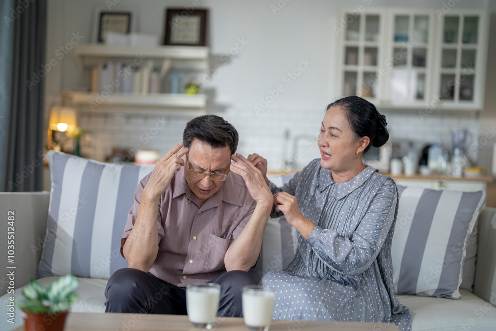 An elderly couple sits on a couch in a cozy living room. The man holds his head in discomfort, while his partner offers support. The image highlights care, concern, and the bond between the couple. An elderly couple sits on a couch in a cozy living room. The man holds his head in discomfort, while his partner offers support. The image highlights care, concern, and the bond between the couple.
