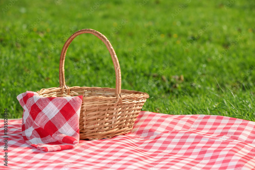 One picnic wicker basket with checkered napkin and blanket on green grass. Space for text One picnic wicker basket with checkered napkin and blanket on green grass. Space for text
