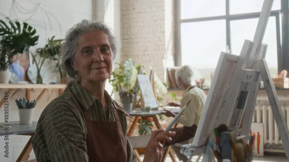 Senior woman in apron painting picture on canvas, then looking at camera in art class, another lady in background Senior woman in apron painting picture on canvas, then looking at camera in art class, another lady in background