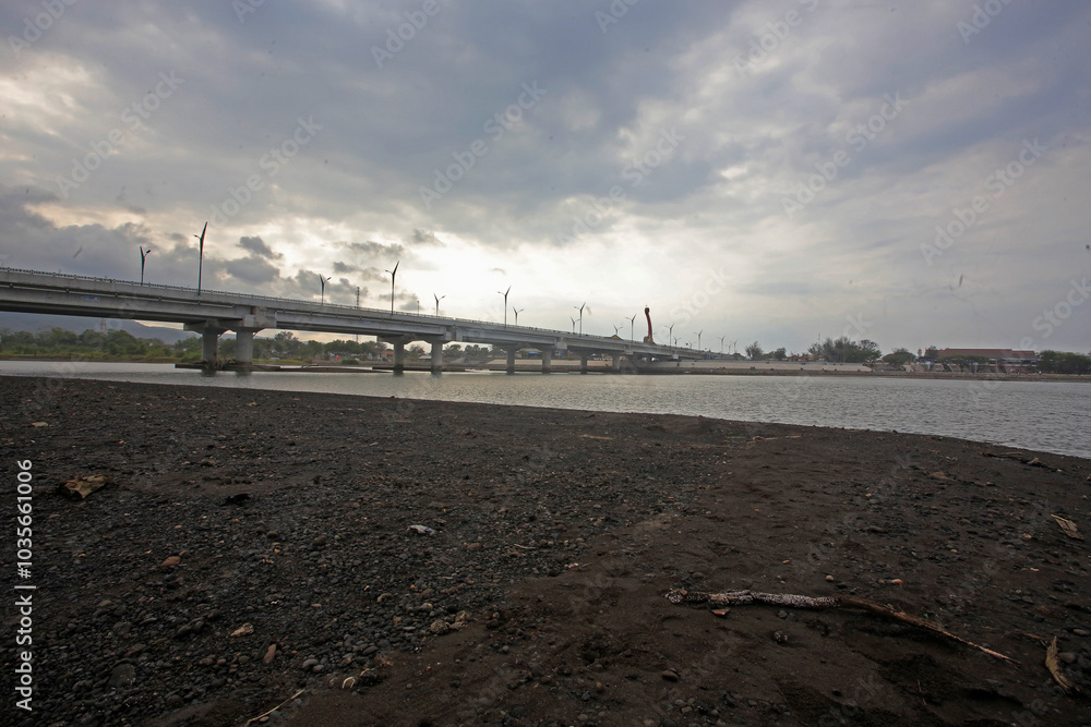 The image shows a large concrete bridge spanning over a river under a cloudy sky, with calm water beneath. Streetlights line the bridge, creating an urban atmosphere around the quiet riverside area The image shows a large concrete bridge spanning over a river under a cloudy sky, with calm water beneath. Streetlights line the bridge, creating an urban atmosphere around the quiet riverside area
