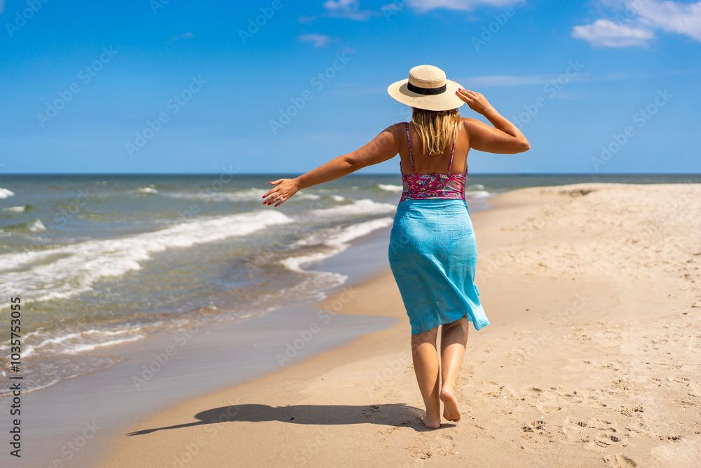 Summer vacation. Tanned woman in colorful swimsuit, blue pareo and sun hat walking by shore on sandy beach on beautiful sunny day. Back view Summer vacation. Tanned woman in colorful swimsuit, blue pareo and sun hat walking by shore on sandy beach on beautiful sunny day. Back view