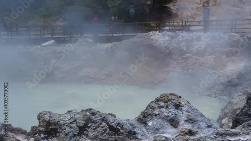 The Sikidang crater in Dieng features a greenish, acidic pool with steam rising from its surface. The surrounding terrain is rocky and barren, indicating the harsh volcanic environment. photo