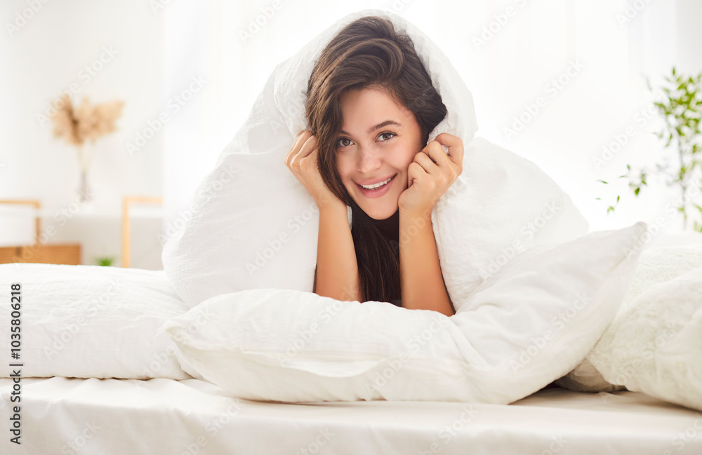 Portrait of happy smiling young woman waking up in bed in white cozy bedroom and looking cheerful at camera covering pillow. Peaceful female person waking up early at home. Good morning concept. Portrait of happy smiling young woman waking up in bed in white cozy bedroom and looking cheerful at camera covering pillow. Peaceful female person waking up early at home. Good morning concept.